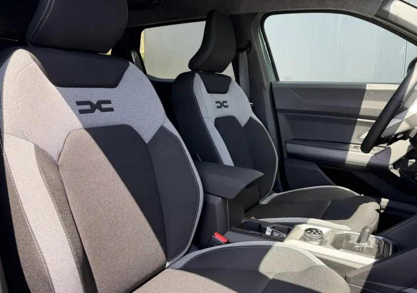 Vue intérieure des sièges avant gris et noir avec logo Dacia, montrant la console centrale et le volant côté droit.