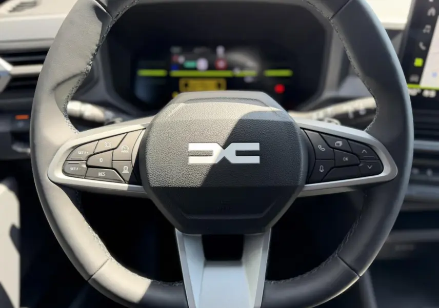 Vue rapprochée du volant noir du Dacia Duster Hybrid-G 150 4x4 avec commandes intégrées et tableau de bord digital flou.