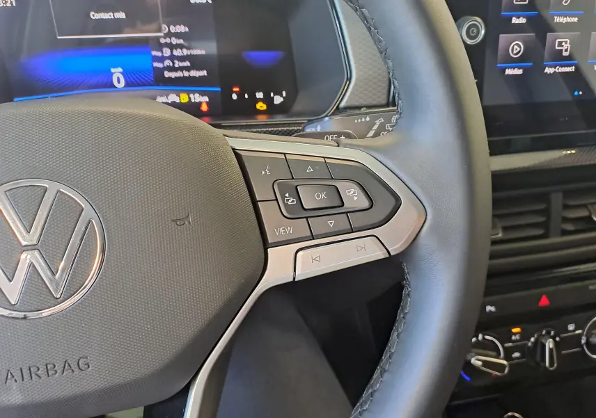 Gros plan sur le volant côté droit du Volkswagen T-Cross 2025 gris cendré, avec commandes multifonctions et tableau de bord numérique.