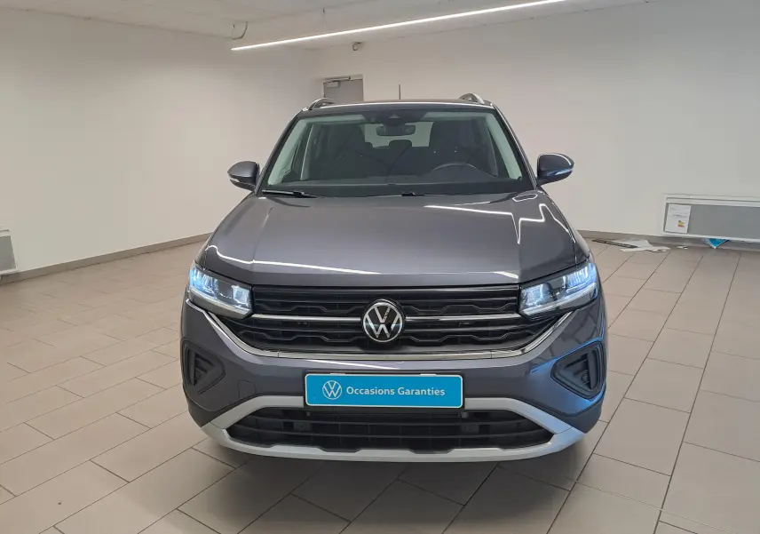 Vue frontale d'un Volkswagen T-Cross 2025 gris cendré avec calandre noire et phares LED allumés en intérieur showroom.