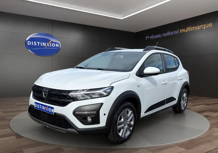 Dacia Sandero Stepway blanc vue 3/4 avant droit, avec barres de toit et protections noires sur les bas de caisse.