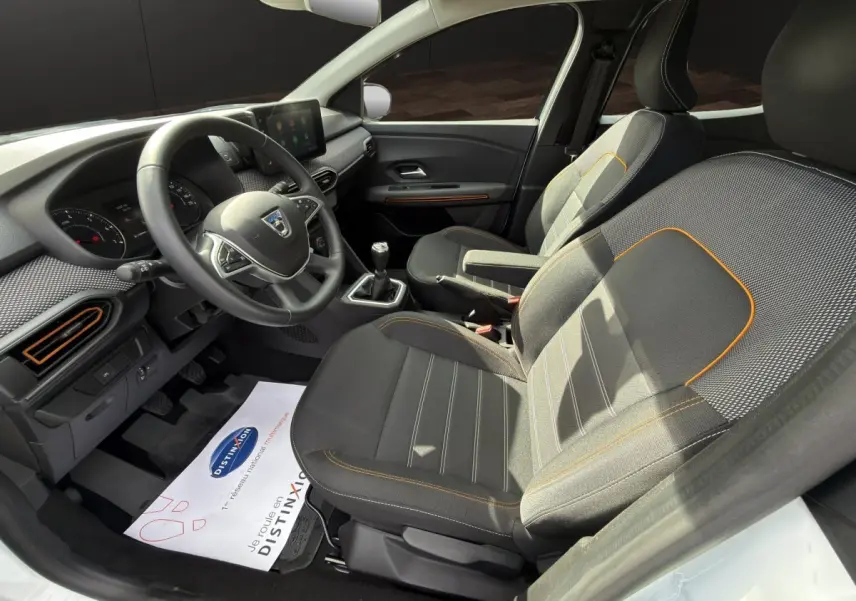Vue intérieure côté conducteur de la Dacia Sandero Stepway blanche, avec volant multifonctions et sièges tissu gris à surpiqûres orange.