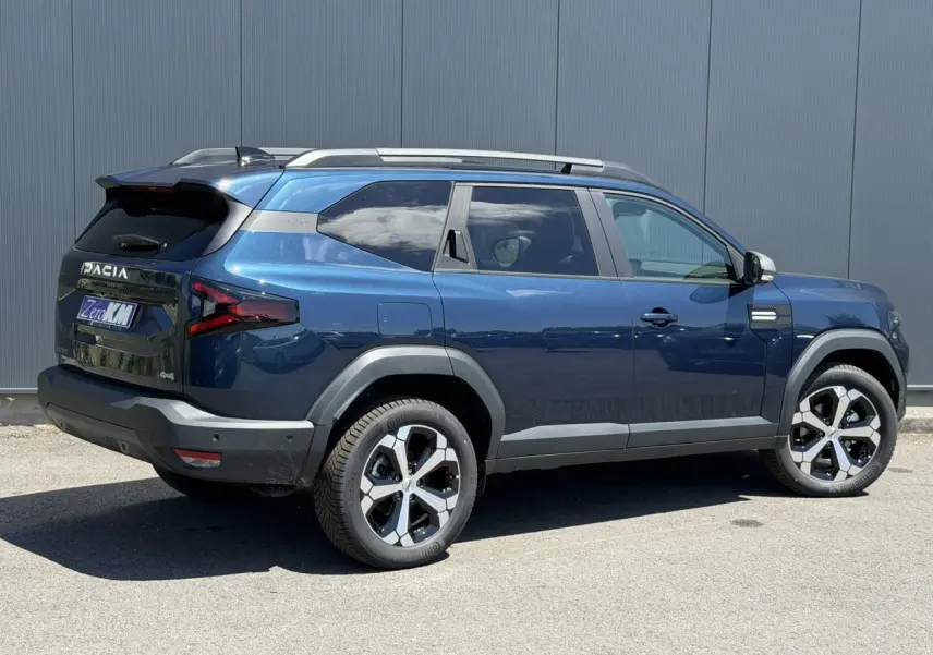 Vue 3/4 arrière droite du SUV Dacia Bigster bleu indigo avec jantes alliage 18 pouces et barres de toit grises.