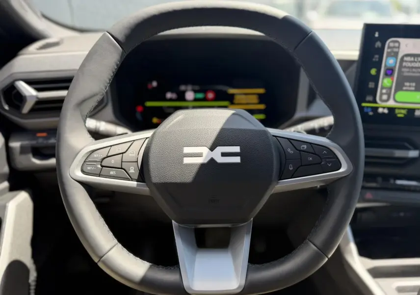Vue rapprochée du volant noir du Dacia Bigster Hybrid-G 150 4X4 avec commandes intégrées et tableau de bord numérique flou.
