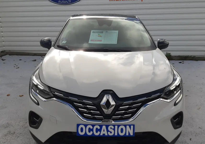 Vue frontale d'un Renault Captur blanc avec toit noir, affichant une plaque "OCCASION" devant un mur publicitaire.