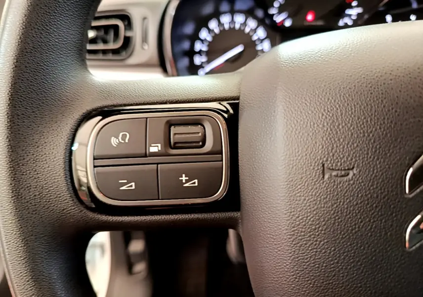 Gros plan sur les commandes gauche du volant noir d'une Citroën C3 blanche, tableau de bord flou en arrière-plan.