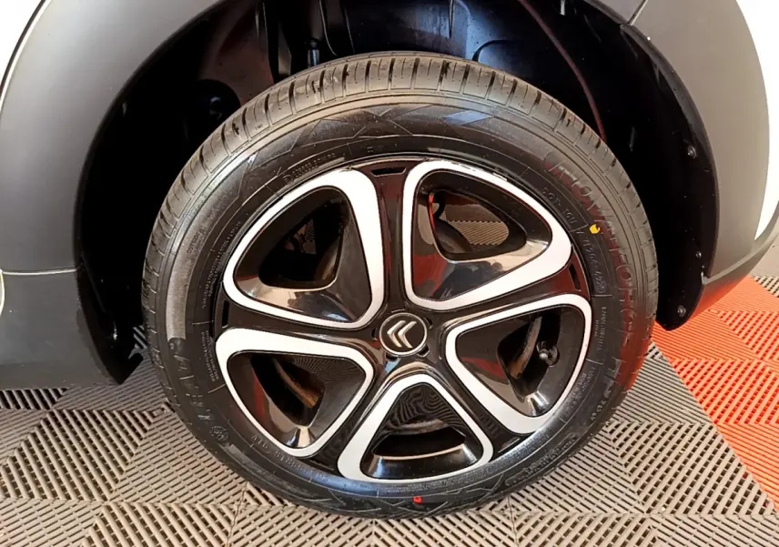 Gros plan sur la roue arrière droite d'une Citroën C3 blanche, avec passage de roue noir et sol carrelé rouge et gris.