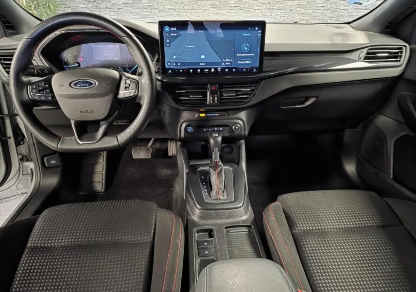 Vue intérieure avant de la Ford Focus 2024 ST Line, tableau de bord noir avec écran tactile central et volant sport à surpiqûres rouges.