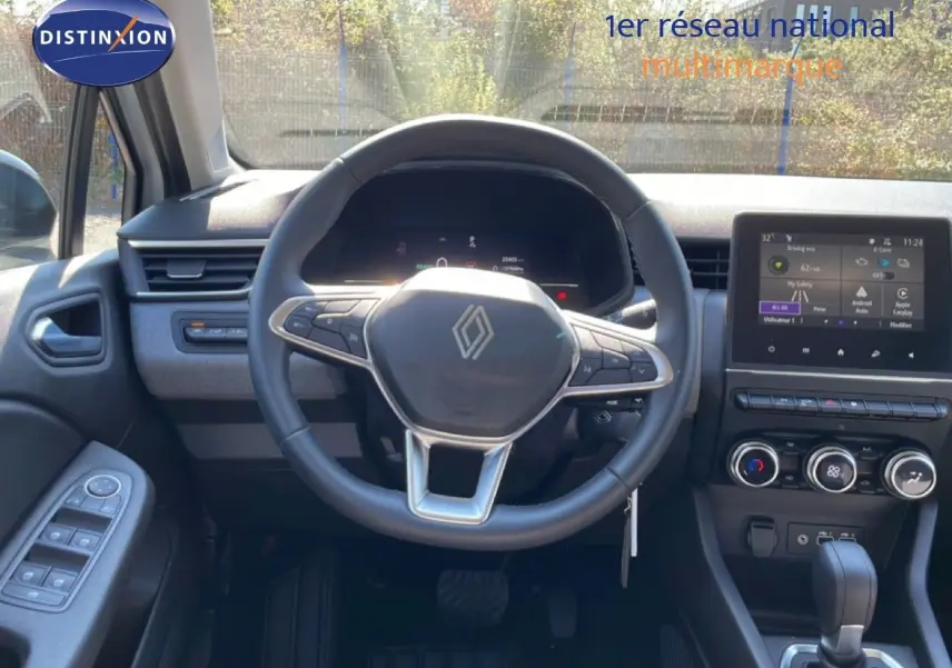 Vue intérieure centrée sur le volant Renault de la Clio blanche, avec tableau de bord numérique et écran tactile visible.