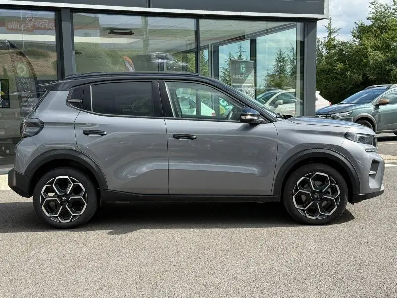 Profil droit d'une Citroën C3 1.2 Turbo 100ch MAX gris Mercury avec jantes alu et toit noir, stationnée devant un bâtiment vitré.
