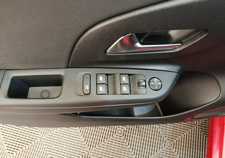 Vue rapprochée de la porte côté gauche de l’Opel Corsa rouge, avec commandes des vitres et poignée intérieure chromée.