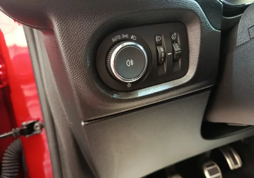 Gros plan sur le panneau de commande des phares à gauche du volant, intérieur noir, portière rouge visible.
