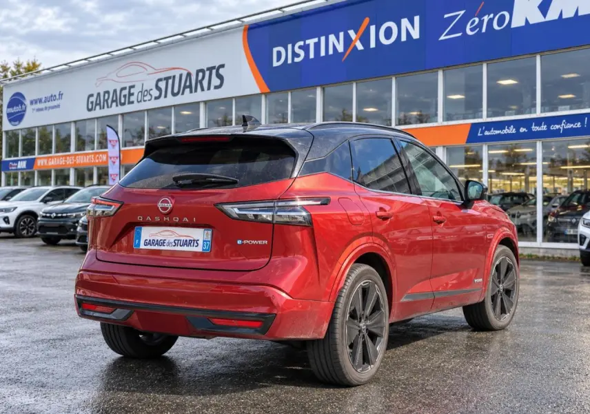 Vue 3/4 arrière d’un Nissan Qashqai 2024 rouge foncé avec toit noir et jantes noires, stationné devant un garage.