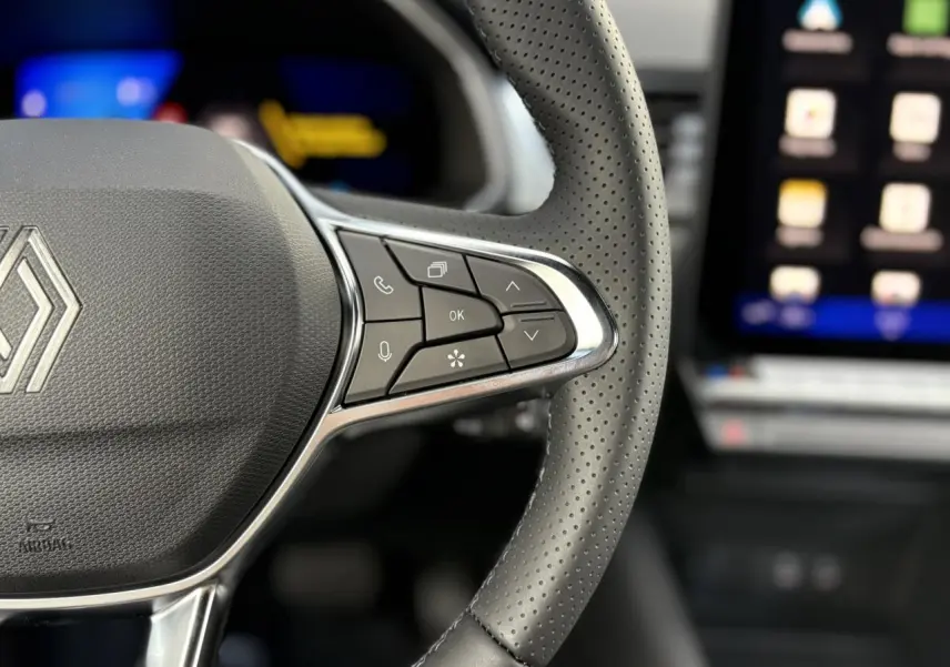 Gros plan sur le volant en cuir perforé du Renault SYMBIOZ 2025 avec commandes multifonctions et écran tactile flou en arrière-plan.