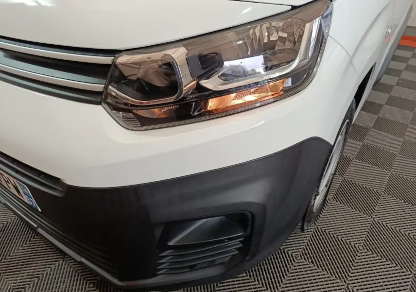 Gros plan sur le phare avant droit allumé du Citroën Berlingo blanc, avec pare-chocs noir et partie de la roue visible.