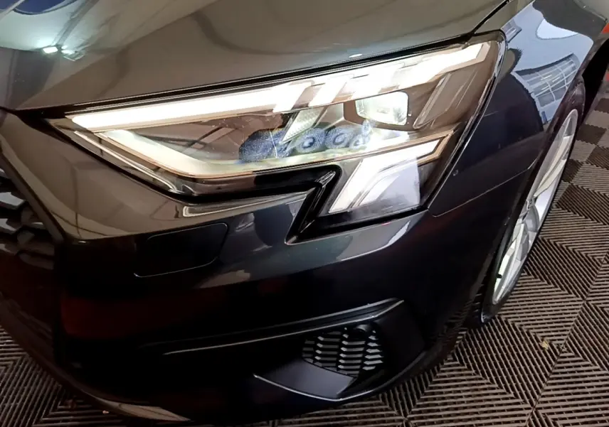 Gros plan sur le phare avant droit LED d’une Audi A3 gris foncé, avec partie de la calandre et roue visible.