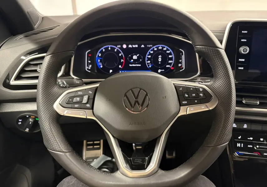 Vue rapprochée du volant noir perforé du Volkswagen T-Roc 1.5 TSI R-Line avec tableau de bord numérique allumé.
