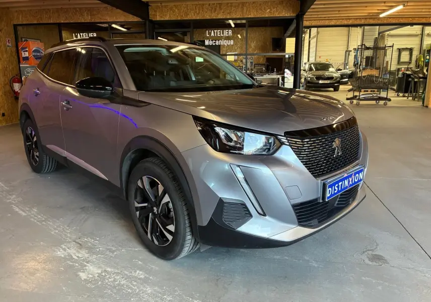 Peugeot 2008 gris en 3/4 avant droit dans un atelier mécanique, avec calandre noire et jantes alliage.