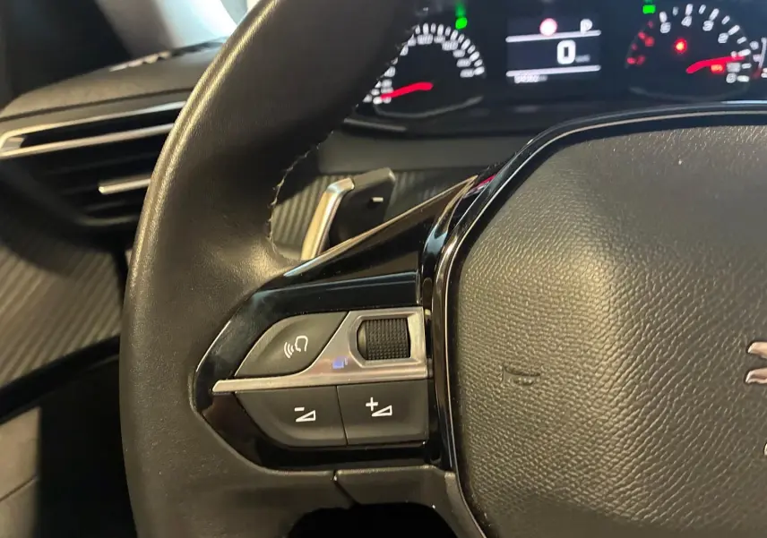 Gros plan sur le volant noir du Peugeot 2008 2022, avec commandes audio et tableau de bord numérique en arrière-plan.