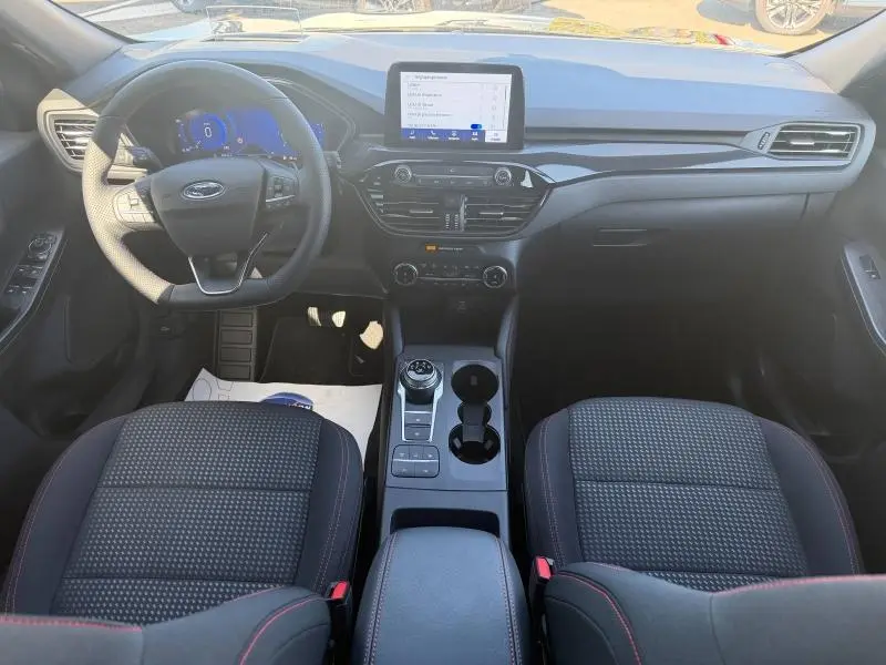 Vue intérieure frontale du Ford Kuga 1.5 EcoBlue 2024, tableau de bord noir avec écran tactile et sièges tissu noir à surpiqûres rouges.