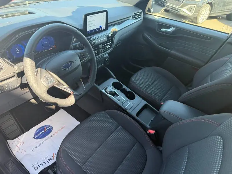Vue intérieure avant droite du Ford Kuga 2024 avec sièges tissu noir, volant cuir multifonction et écran tactile central.