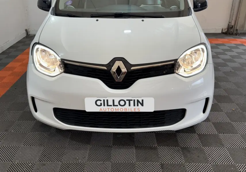 Vue frontale rapprochée de la Renault Twingo blanche avec phares allumés dans un garage.