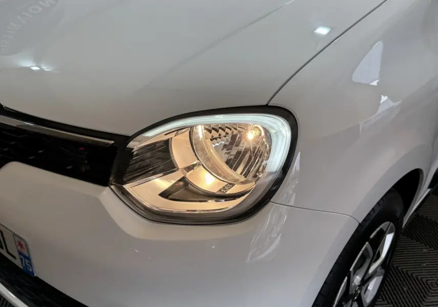 Gros plan sur le phare avant droit allumé d'une Renault Twingo blanche 2022, avec partie de la calandre visible.