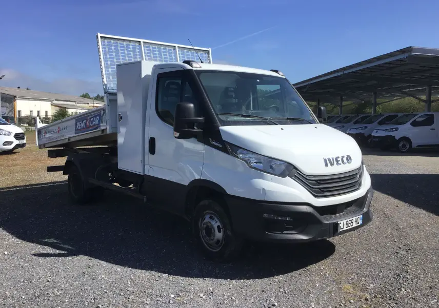 Vue 3/4 avant droite d’un Iveco Daily blanc avec benne acier et coffre latéral, stationné sur un sol gravillonné.