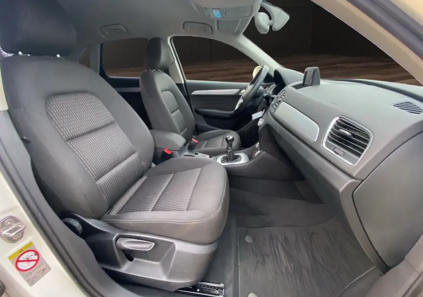 Vue intérieure côté conducteur de l'Audi Q3 blanc, montrant les sièges noirs en tissu et la console avec GPS intégré.