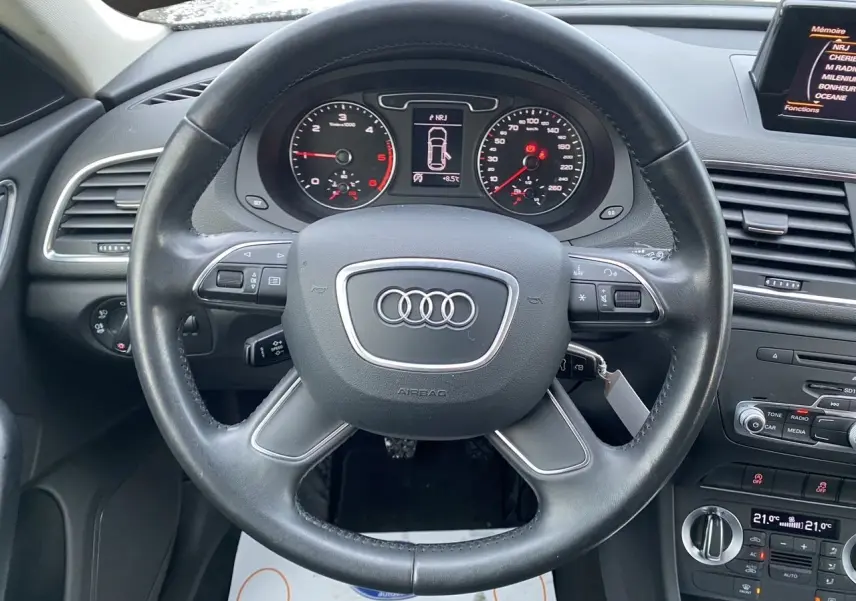 Vue intérieure centrée sur le volant cuir noir d’un Audi Q3 blanc de 2014 avec tableau de bord et écran GPS visible.