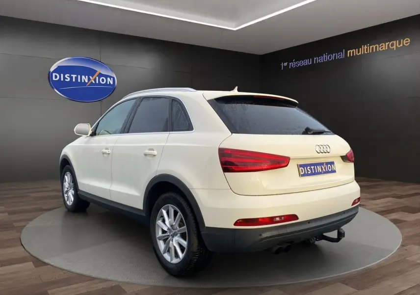 Audi Q3 blanc en 3/4 arrière droit, vitres arrière surteintées et attelage visibles dans un showroom.