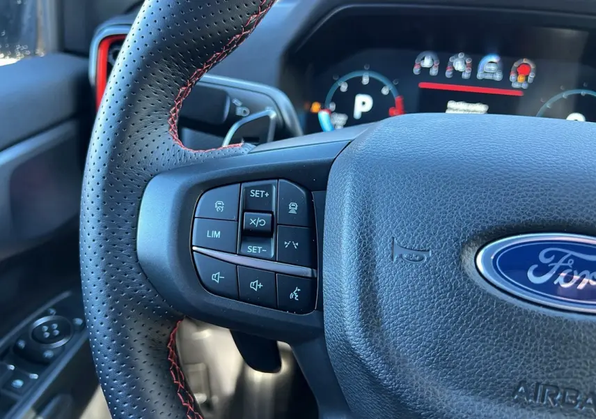 Gros plan sur le volant cuir perforé noir avec surpiqûres rouges du Ford Ranger Raptor 2025, boutons multifonctions visibles.