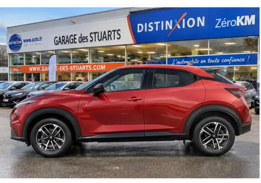 Vue latérale gauche d'un Nissan Juke 1.6 Hybrid rouge Fuji Sunset avec jantes alliage 17 pouces, garé devant un garage.