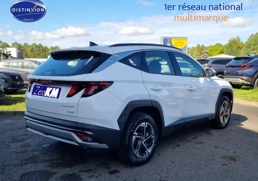 Vue 3/4 arrière droite du Hyundai Tucson blanc atlas hybride avec jantes alliage et barres de toit visibles.