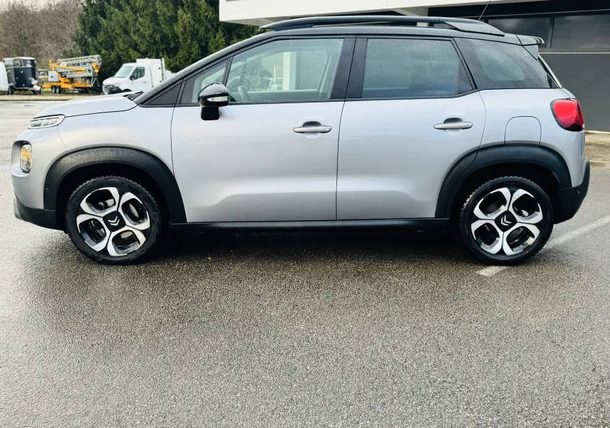 Profil côté gauche d'une Citroën C3 Aircross gris Artense avec toit noir et protections noires sur un sol humide.