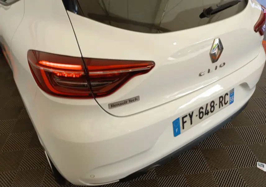 Vue 3/4 arrière droite d'une Renault Clio blanche avec feux LED allumés et badge Renault Tech visible sur le coffre.