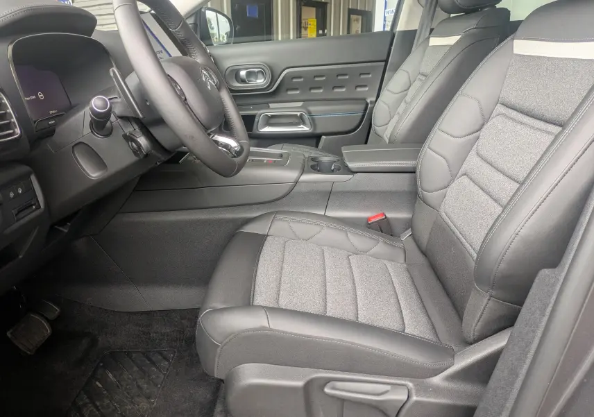 Vue intérieure côté conducteur du Citroën C5 Aircross hybride 2025, sièges tissu/TEP gris avec volant cuir et console centrale.