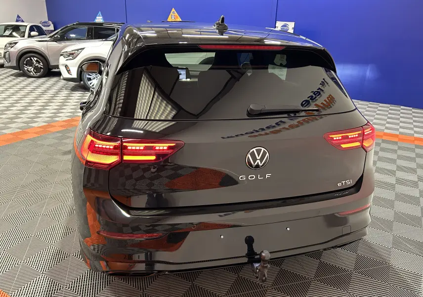 Vue arrière d'une Volkswagen Golf VIII eTSI noire métal avec feux arrière allumés et attelage visible en intérieur showroom.