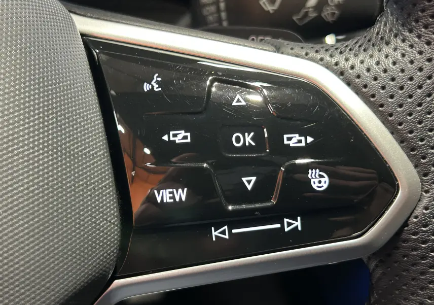 Gros plan sur les commandes multifonctions noires du volant perforé d'une Volkswagen Golf VIII eTSI noire, boutons rétroéclairés blancs.