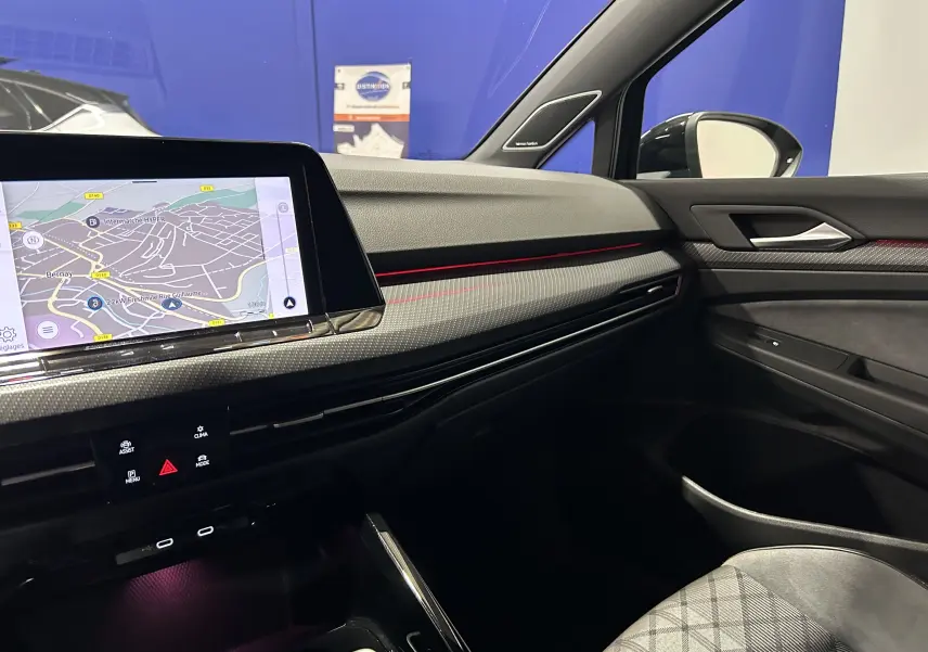 Vue intérieure côté passager de la Volkswagen Golf VIII eTSI noire, avec écran GPS et bandeau lumineux rouge sur la planche de bord.
