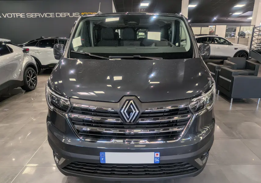Vue frontale d'un Renault Trafic Combi gris Comète 2025 en showroom, avec calandre chromée et logo Renault bien visible.