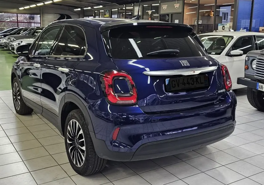 Vue 3/4 arrière d'un FIAT 500X bleu 2024 hybride essence en showroom, avec feux arrière distinctifs et jantes stylées.
