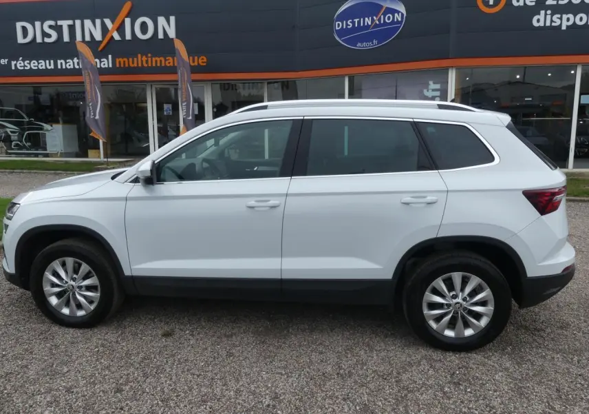 Profil côté gauche d'un SUV SKODA KAROQ blanc 2024 avec jantes alliage 17 pouces devant un concessionnaire Distinxion.