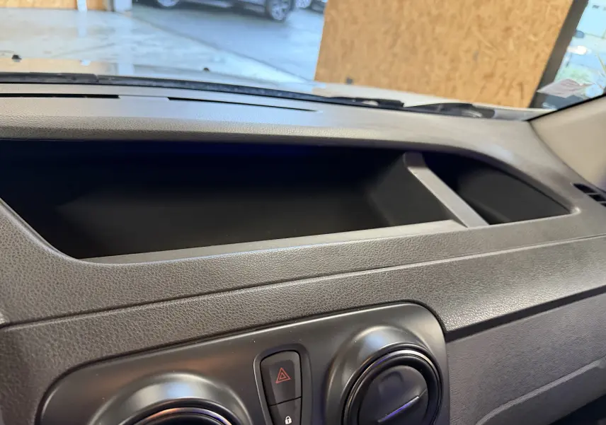 Détail du tableau de bord noir du Dacia Dokker gris, montrant les commandes de ventilation et le rangement supérieur.