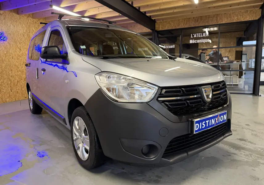 Vue 3/4 avant droit d'un Dacia Dokker gris 2019 avec pare-chocs noir mat et calandre distinctive en intérieur d'atelier.