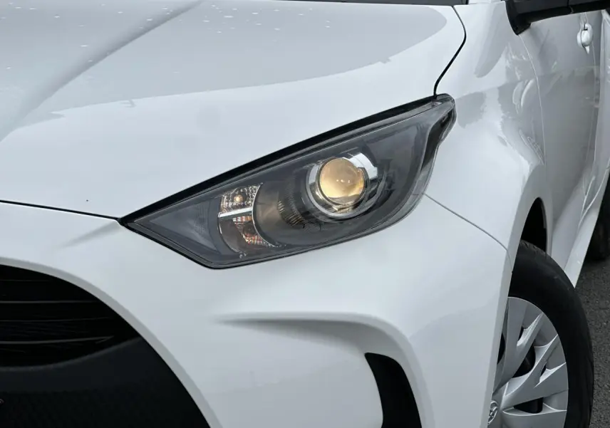Gros plan sur le phare avant droit allumé d'une Toyota Yaris hybride blanche 2025, avec jante et aile visibles.