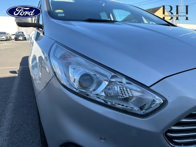Gros plan sur le phare avant droit d’un Ford S-MAX gris lunaire, avec calandre chromée visible en arrière-plan.