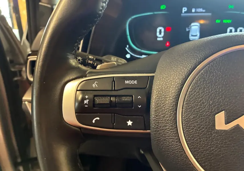Gros plan sur les commandes gauche du volant du Kia Sportage hybride gris 2024 avec tableau de bord numérique en arrière-plan.