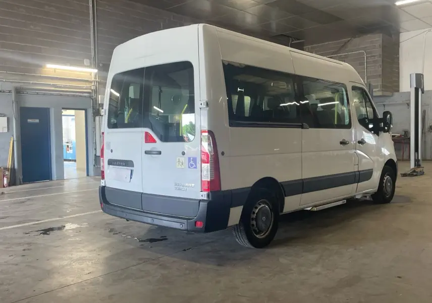 Renault Master blanc en 3/4 arrière droit dans un garage, avec rampe arrière et logo handicap visible sur la porte.