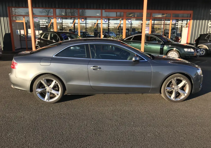 Audi A5 Coupé gris dauphin vu de profil côté gauche, avec jantes alliage 5 branches et silhouette élégante.
